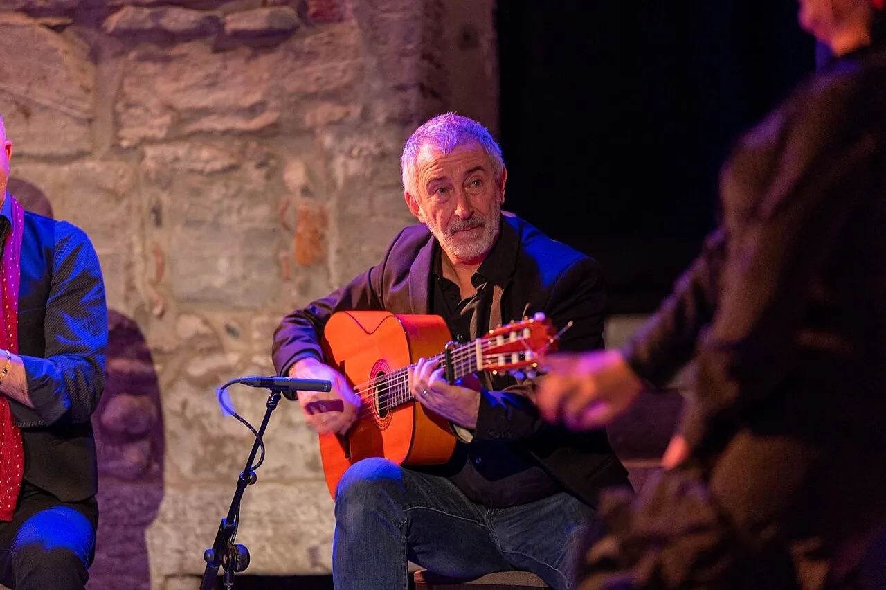 En flamencoguitarist i en mørk jakke sidder under scenelyset og spiller på en guitar med orange top, med en stenmur bag sig