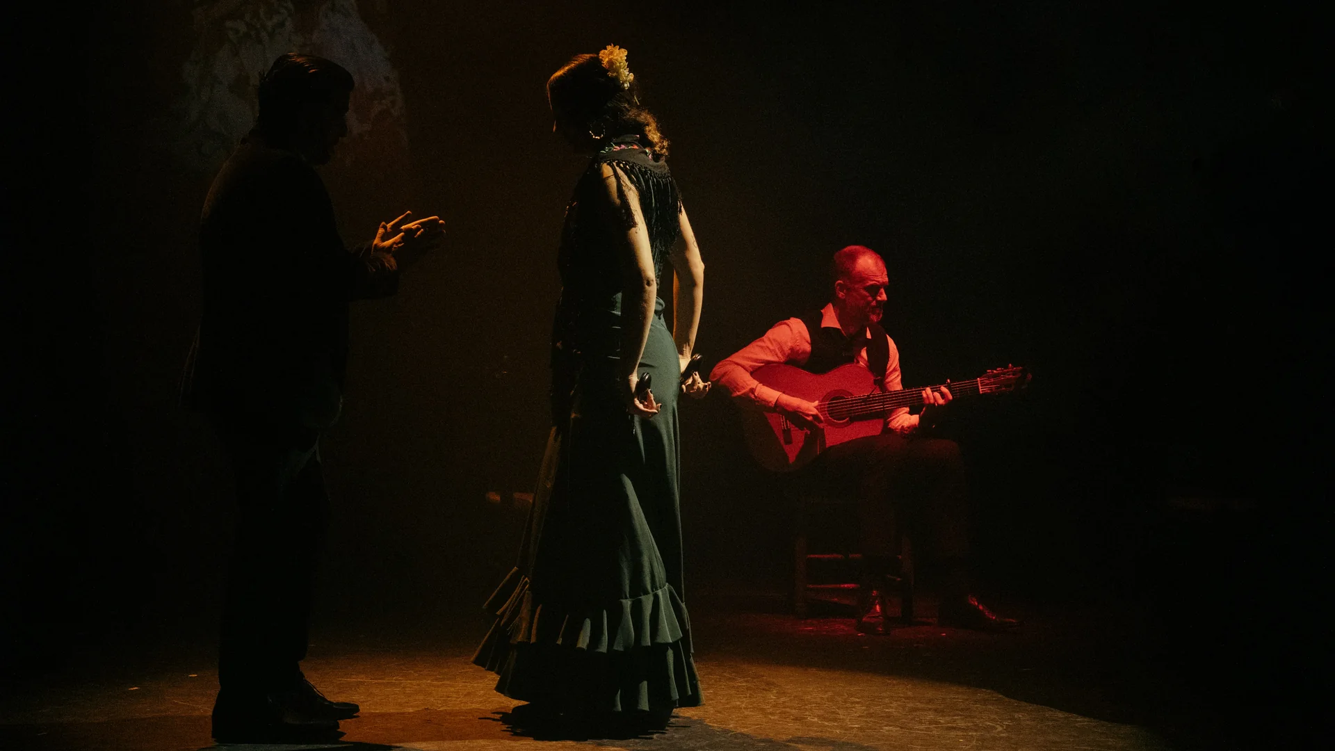 En flamencodanser i mørkegrøn kjole med flæser står i varmt scenelys ved siden af en siddende guitarist og en sanger i silhuet på en dæmpet scene