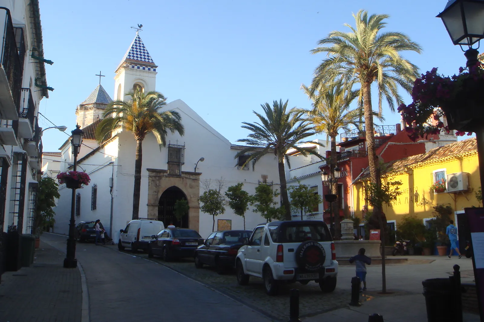 En hvidkalket kirke med et lille tårn på en stille plads i Marbella, omgivet af palmer, parkerede biler og et gult hus ved siden af